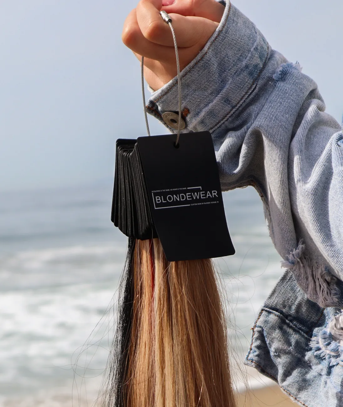 A BlondeWear swatch ring held up at the beach, long blonde and brunette samples hanging from it