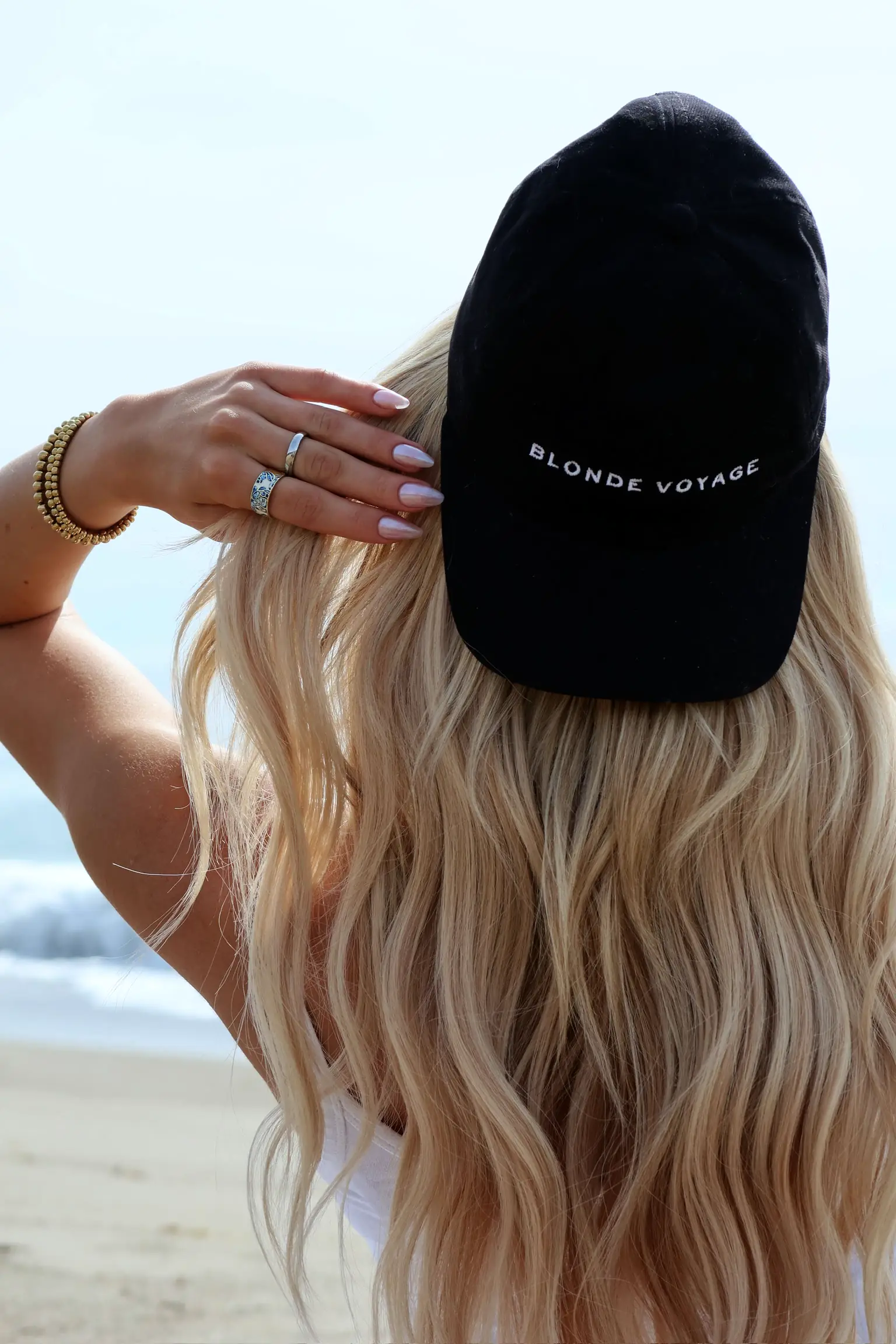 Long beachy blonde waves under a black Blonde Voyage cap, at the shoreline