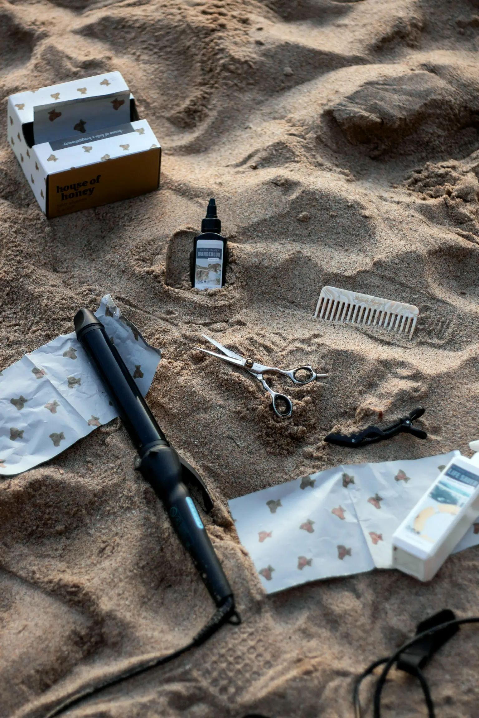 A curling iron, shears, a comb and TRVL bottles laid out on beach sand